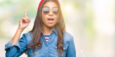 Genç güzel Arap kadın başarılı fikirle izole arka plan işaret parmağı üzerinde güneş gözlüğü takıyor. Çıktıktan ve mutlu. Bir numara.