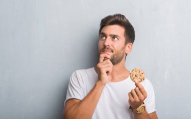 Handsome young man over grey grunge wall eating chocolate chip cooky serious face thinking about question, very confused idea clipart