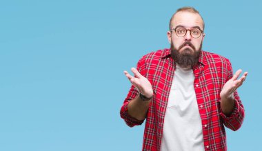 Young caucasian hipster man wearing glasses over isolated background clueless and confused expression with arms and hands raised. Doubt concept. clipart