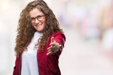 Güzel esmer kıvırcık saçlı Genç kız dostu sunan el sıkışma tebrik ve davet edici olarak gülümseyen izole arka plan üzerinde ceket ve gözlük takıyor. Başarılı iş.