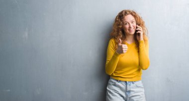 Genç Kızıl saçlı kadın büyük memnun Evet gri grunge duvarın üzerinden gülümseme ok işareti, başparmak ile parmak, mükemmel işareti yapıyor