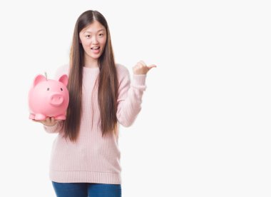 İşaret ve başparmak kadar mutlu yüz gülümseyen tarafı ile gösterilen kumbara tutan izole arka plan üzerinde Genç Çinli kadın