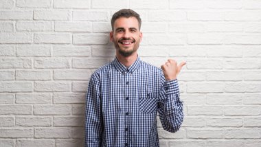 Genç yetişkin adam işaret ve başparmak kadar mutlu yüz gülümseyen tarafı ile gösterilen beyaz tuğla duvarın üzerinden ayakta