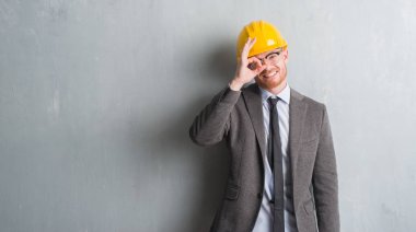 Mimar kıyafeti giymiş genç Kızıl saçlı adam ve ok işareti ile el parmaklarının arasından seyir göze yapıyor gülümseyen mutlu yüz ile helment