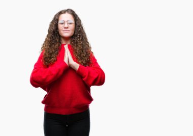 Ellerle birlikte AF emin gülümseyen için soran dua izole arka plan üzerinde gözlük ve kış kazak giyen güzel esmer kıvırcık saçlı Genç kız.