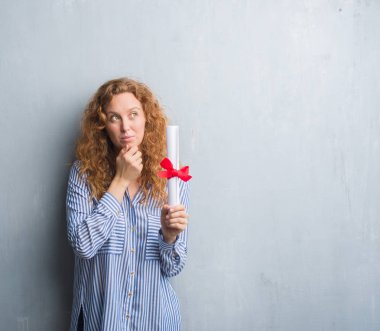 Genç redhead iş kadını diploma ciddi bir yüz düşünme hakkında soru, çok karışık fikir tutan gri grunge duvarın üzerinden