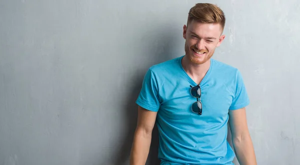 Young redhead man over grey grunge wall wearing casual outfit winking looking at the camera with sexy expression, cheerful and happy face.