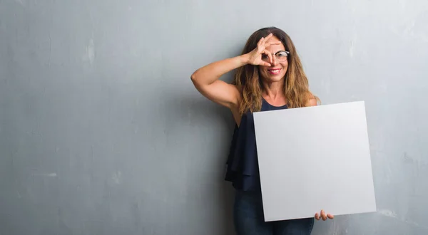 Middle age hispanic woman standing over grey grunge wall holding blank banner with happy face smiling doing ok sign with hand on eye looking through fingers