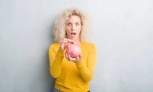 Young Blonde Woman Grunge Grey Background Holding Piggy Bank Scared — Stock Photo, Image