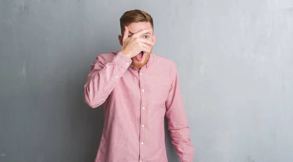 Young redhead man over grey grunge wall wearing pink shirt peeking in shock covering face and eyes with hand, looking through fingers with embarrassed expression.