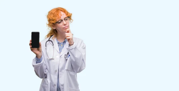 Young Redhead Doctor Woman Showing Smartphone Serious Face Thinking Question — Stock Photo, Image