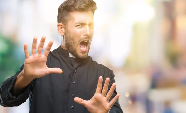 Young catholic christian priest man over isolated background afraid and terrified with fear expression stop gesture with hands, shouting in shock. Panic concept.