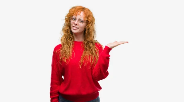 陽気な提示とカメラを見て手の手のひらで指している笑みを浮かべて赤いセーターを着ている若い赤毛の女性 — ストック写真