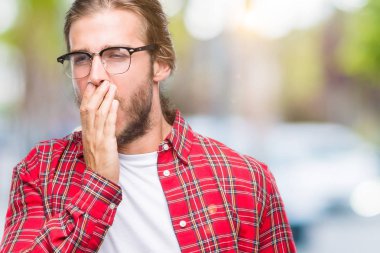 Genç yakışıklı adam uzun saçlı gözlüklü izole arka plan üzerinde el ile ağız kapsayan yorgun esneme sıkılmış. Restless ve uyku hali.