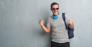 Genç beyaz öğrenci adam gri grunge duvar giyen kulaklık ve gurur çığlık ve zafer ve başarı çok heyecanlı, kutluyor sırt çantası duygu tezahürat