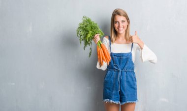 Güzel genç kadın taze havuç ile mutlu büyük holding grunge gri duvar yüzünden gülümseme ok işareti, başparmak ile parmak, mükemmel işareti yapıyor
