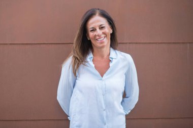 Güzel orta yaş İspanyol kadın ayakta ve diş gösterilen kendine güvenen bir gülümsemeyle gülümseyerek mutlu bir yüz ile güneşli bir tuğla duvarın üzerinden