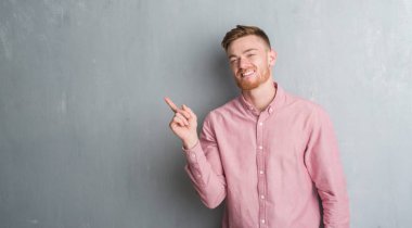 Genç redhead gri grunge duvar giyen üzerinde pembe gömlek uzun kameraya bakarak yan el ve parmak ile işaret yüzünde kocaman bir gülümseme ile.