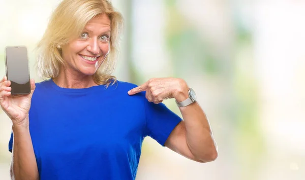 Middle age blonde woman showing blank screen of smartphone over isolated background with surprise face pointing finger to himself