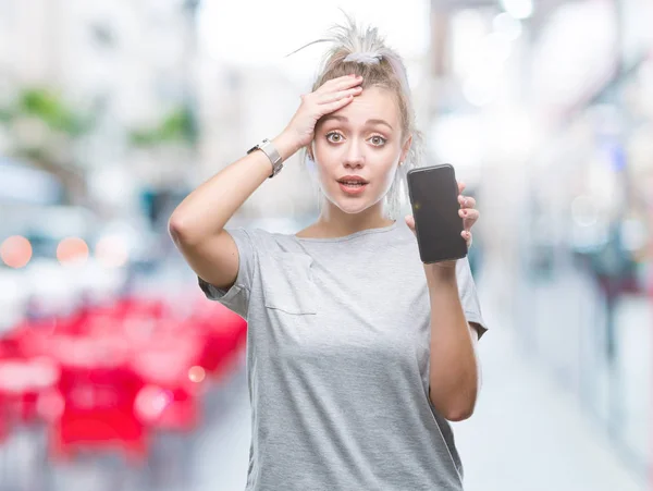 Young blonde woman showing screen of smartphone over isolated background stressed with hand on head, shocked with shame and surprise face, angry and frustrated. Fear and upset for mistake.