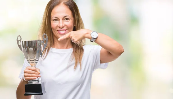 Middle age hispanic winner woman celebrating award holding trophy over isolated background very happy pointing with hand and finger