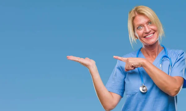 中年金发护士外科医生妇女在孤立背景惊讶和微笑的相机 而提出与手和手指指向 — 图库照片
