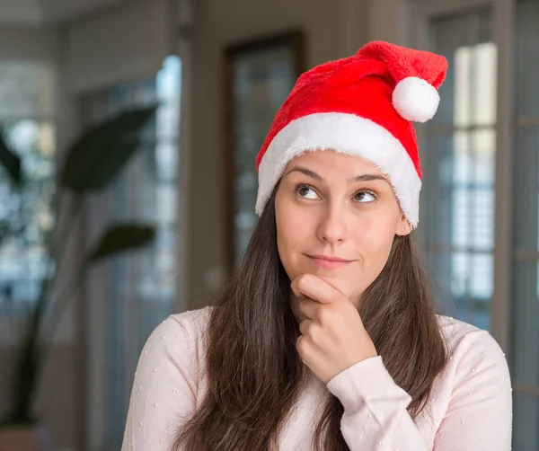 美丽的年轻女子戴着圣诞老人的帽子在家里看着自信的镜头与交叉的手臂和手提出的下巴上的微笑 积极的思考 — 图库照片