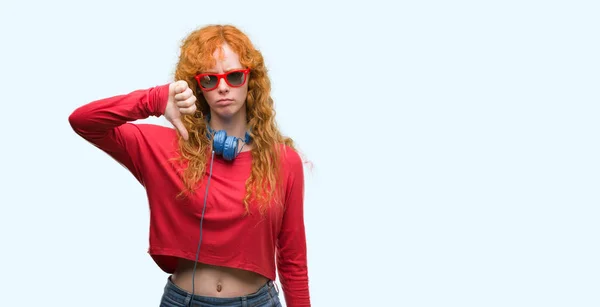 年轻的红发女郎戴着耳机与愤怒的脸 阴性符号显示不喜欢拇指向下 拒绝概念 — 图库照片