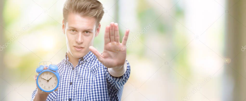 Young handsome blond man holding alarm clock with open hand doing stop sign with serious and confident expression, defense gesture