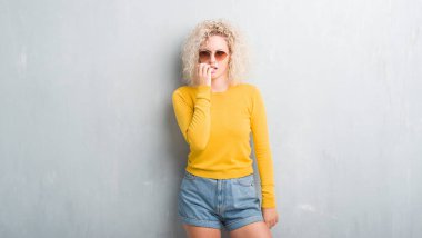 Stresli ve gergin elle çivi ısırma ağzına bakarak grunge gri arka plan üzerinde Kıvırcık saçlı sarışın genç kadın. Anksiyete sorunu.