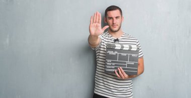 Beyaz delikanlı film fıçı tahtası ile tutarak gri grunge duvarın üzerinden açık el dur işareti ile ciddi ve kendine güvenen ifade, savunma hareketi yapıyor