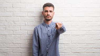 Genç yetişkin adam kamera ve size, el işareti, pozitif ve kendinden emin bir jest ön parmak ile işaret beyaz tuğla duvarın üzerinden ayakta