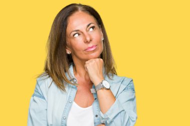 Orta yaş yetişkin kadın elle çene düşünme hakkında soru, dalgın ifade izole arka plan üzerinde rahat kot gömlek giymiş. Düşünceli yüzüyle gülümsüyor. Şüphe kavramı.