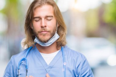 Göğüs kapalı gözlerle ellerinde ve yüzünde minnettar jest ile gülümseyen izole arka plan üzerinde uzun saçlı Genç yakışıklı Doktor adam. Sağlık kavramı.