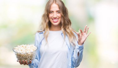 Güzel genç sarışın kadın ayakta ve diş gösterilen kendine güvenen bir gülümsemeyle gülümseyerek mutlu bir yüz ile izole arka plan üzerinde patlamış mısır yemek