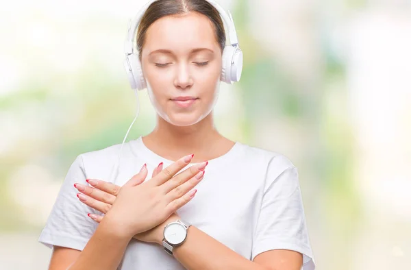 年轻的白种人妇女听音乐戴着耳机在孤立的背景微笑与手在胸部与紧闭的眼睛和感恩的手势在脸上 健康理念 — 图库照片