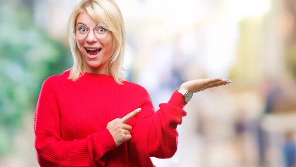驚くし 手とを提示 指で指している間カメラに笑顔孤立の背景にセーターを着て メガネを着て若い美しいブロンドの女性 — ストック写真