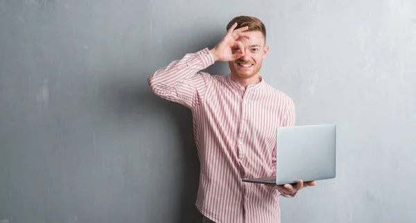 Young redhead man over grey grunge wall holding and using computer laptop with happy face smiling doing ok sign with hand on eye looking through fingers