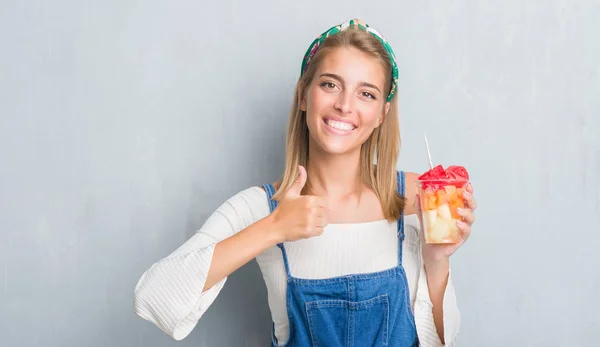 美丽的年轻女人超过垃圾灰墙吃水果高兴与大微笑做 拇指与手指 优秀的标志 — 图库照片