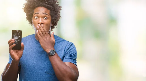 Afro american man holding broken smartphone over isolated background cover mouth with hand shocked with shame for mistake, expression of fear, scared in silence, secret concept