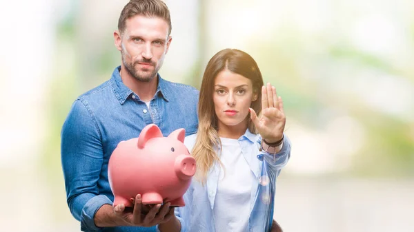 Young couple in love holding piggy bank over isolated background with open hand doing stop sign with serious and confident expression, defense gesture