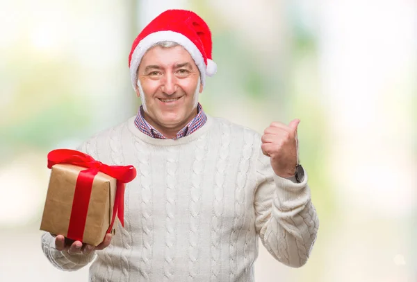 英俊的老人戴着圣诞帽子和持有的礼物 在孤立的背景指向和显示与拇指面带笑脸微笑 — 图库照片