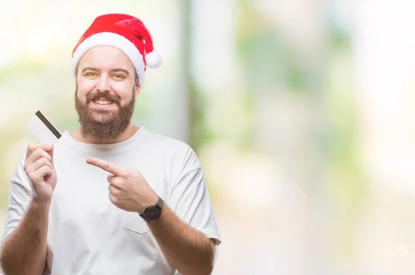 若い白人男の手と指で指している非常に幸せの分離の背景の上クレジット カードを保持しているクリスマス帽子をかぶっています — ストック写真