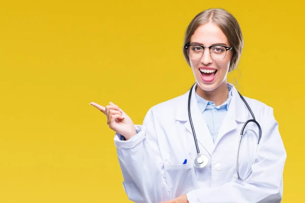 Beautiful young blonde doctor woman wearing medical uniform over isolated background with a big smile on face, pointing with hand and finger to the side looking at the camera.