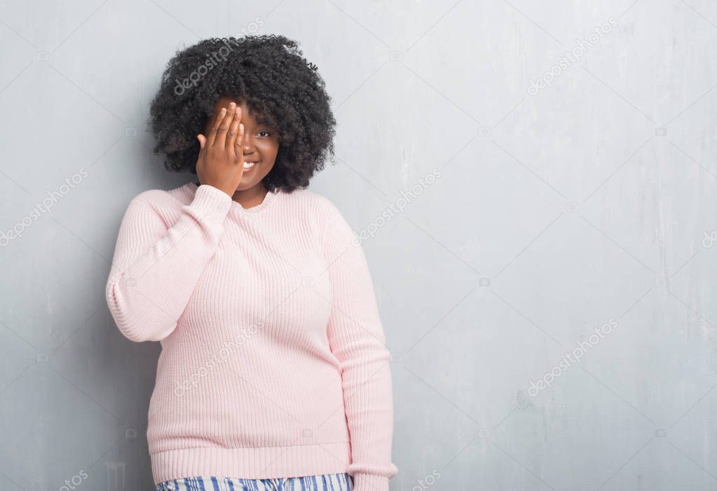 Young african american plus size woman over grey grunge wall wearing winter sweater covering one eye with hand with confident smile on face and surprise emotion.