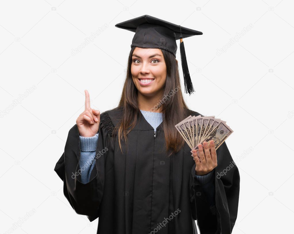 Young hispanic woman wearing graduated uniform holding dollars surprised with an idea or question pointing finger with happy face, number one