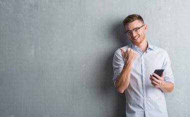 Genç Kızıl saçlı adam işaret ve başparmak kadar mutlu yüz gülümseyen tarafı ile gösterilen smartphone kullanarak ileti gönderme gri grunge duvarın üzerinden