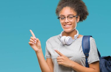 Gülümseyen ve iki elleri ve parmakları ile tarafına işaret kamera bakarak izole arka plan üzerinde kulaklık ve sırt çantası giyen genç afro Amerikan öğrenci kadın.