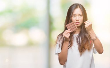 Genç Asyalı kadın çikolata üzerinde izole yeme arka plan ile öfke, çılgın ve yükseltilmiş eliyle diye bağıran rahatsız ve hayal kırıklığına uğramış öfke kavramı bağırarak