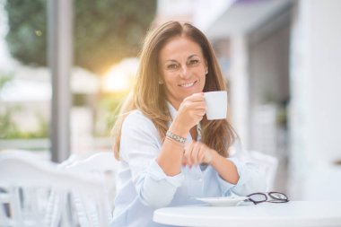 Beautiful middle age hispanic woman at cafeteria with smile on face at the town. Drinking cup of coffee confident and cheerful at the terrace on a sunny day. clipart